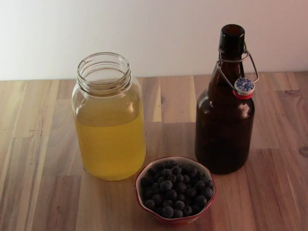 a bowl of blueberries with a mason jar of kombucha and an emply swing top bottle