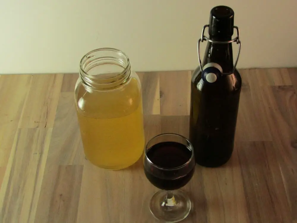 A glass of grape juice a mason jar with kombucha and an empty swing top bottle