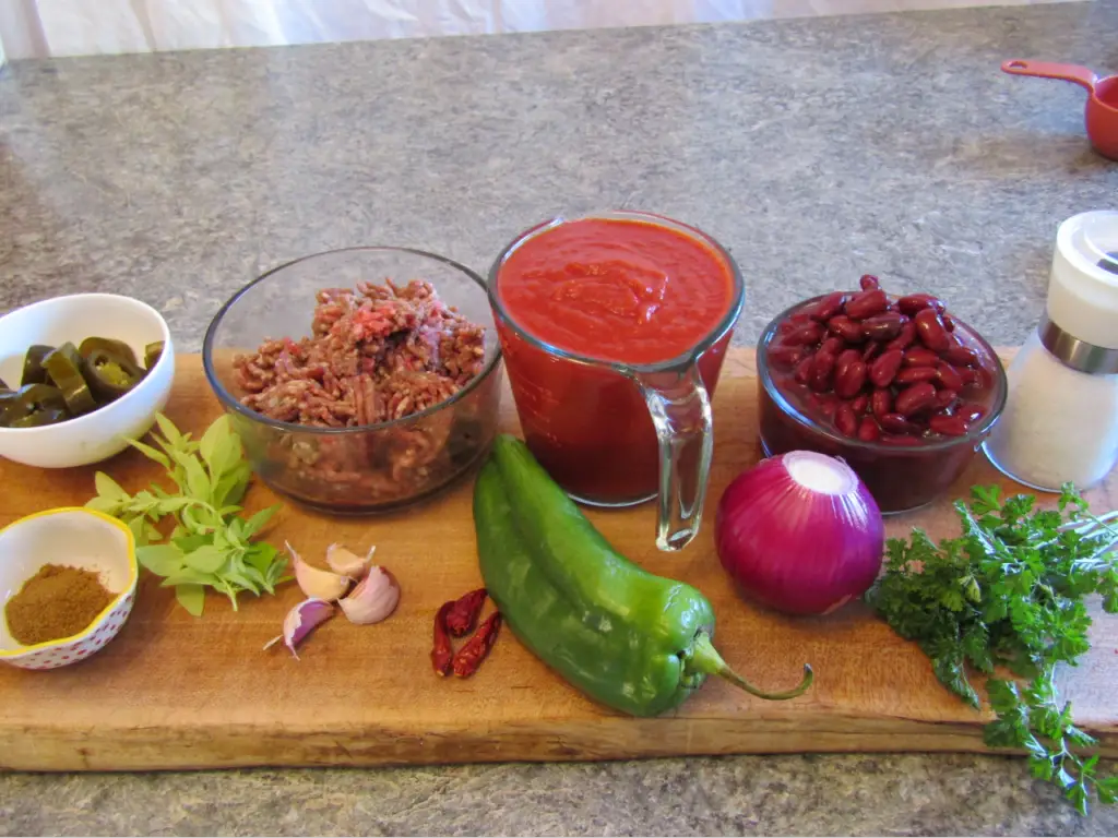 Ingredients for chili with fermented hot peppers: ground beef, tomaoto sauce, kidney beens, onion, garlic, fermented jalepeno peppers fresh oragano and parsly, bowl of cumin, salt and 3 hot chili peppers