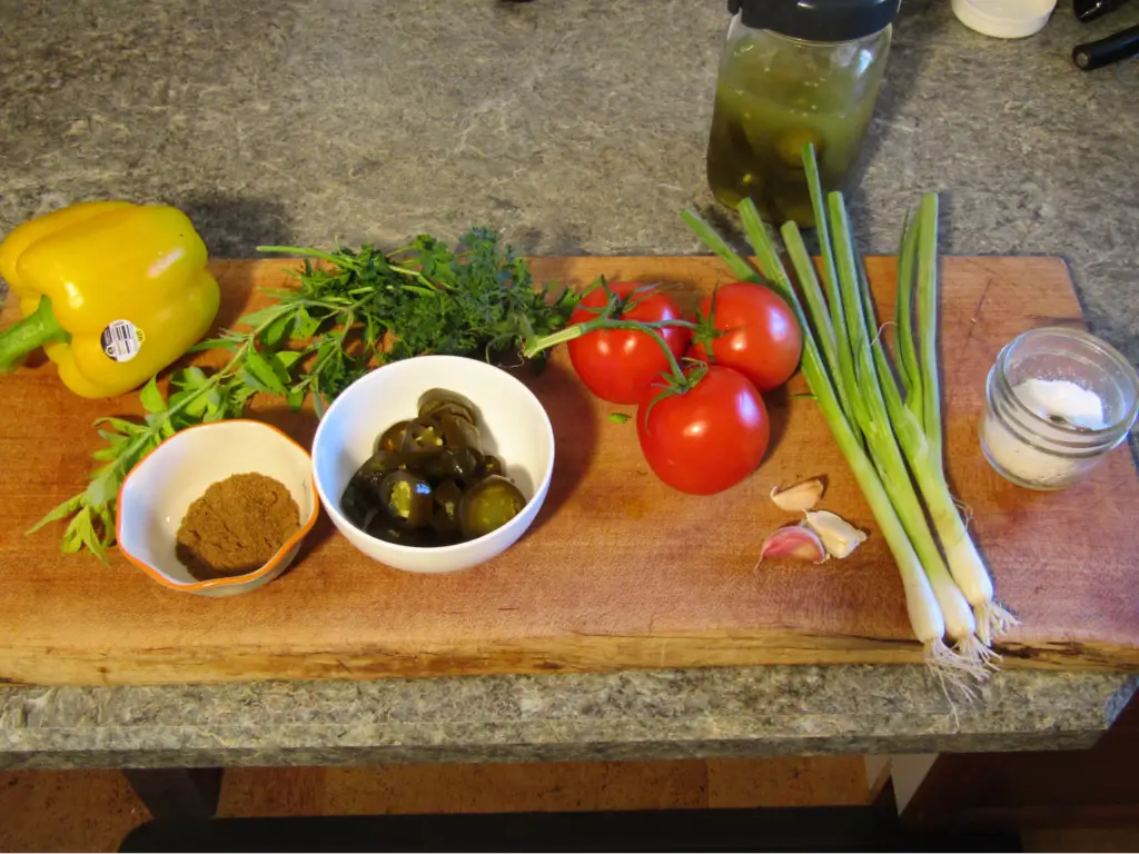 Ingredients for fresh salsa made with fermented hot peppers: tomatos, bell pepper, green onions, fermented peppers in a bowl, garlic, fresh oragano and parsly, powdered cumin in a bowl and a container of salt