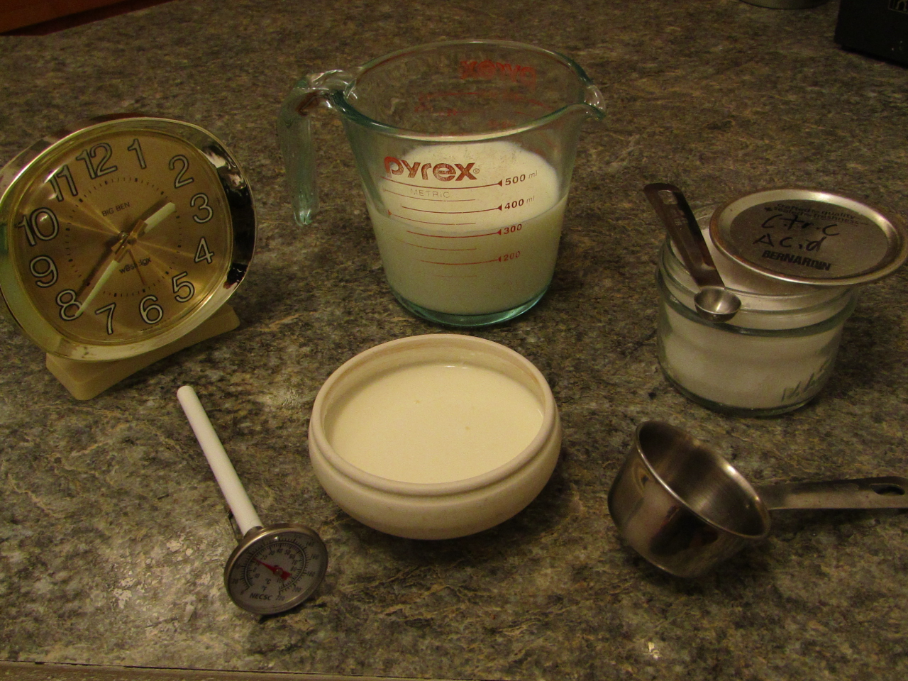A bowl of yogurt with a thermoneter, clock measuring cup with milk, jar with citric acid and a measuring cup surrounding it.