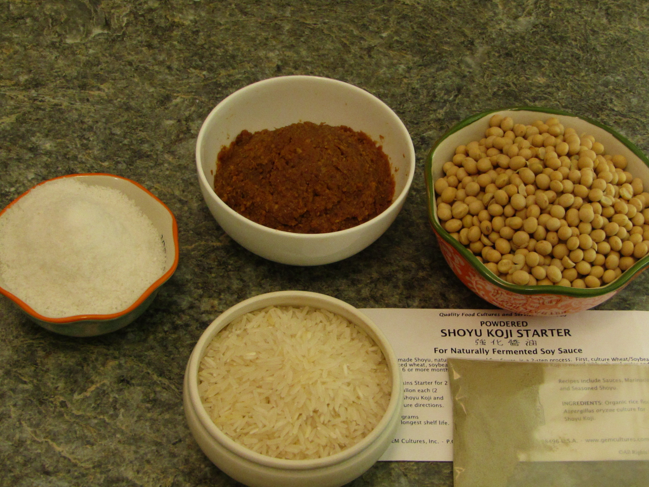 four bowls of various sizes, one containing miso, one soy beans, one filled with salt and another containing rice with a packet of kogi mold