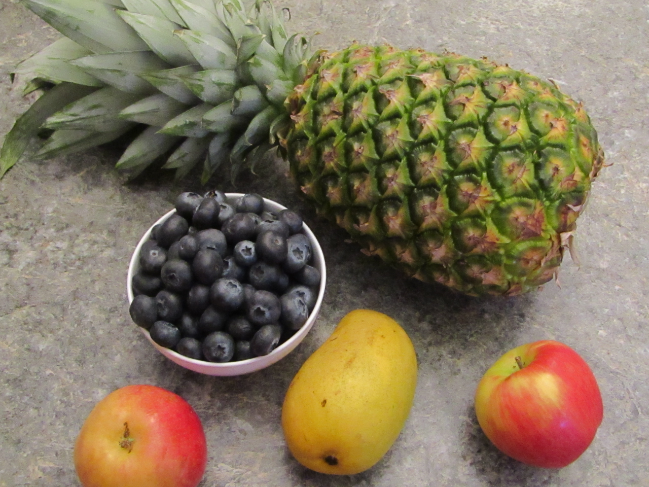 An arraingement of pineapple, blueberries in a bowl, two apples and a mango on a counter